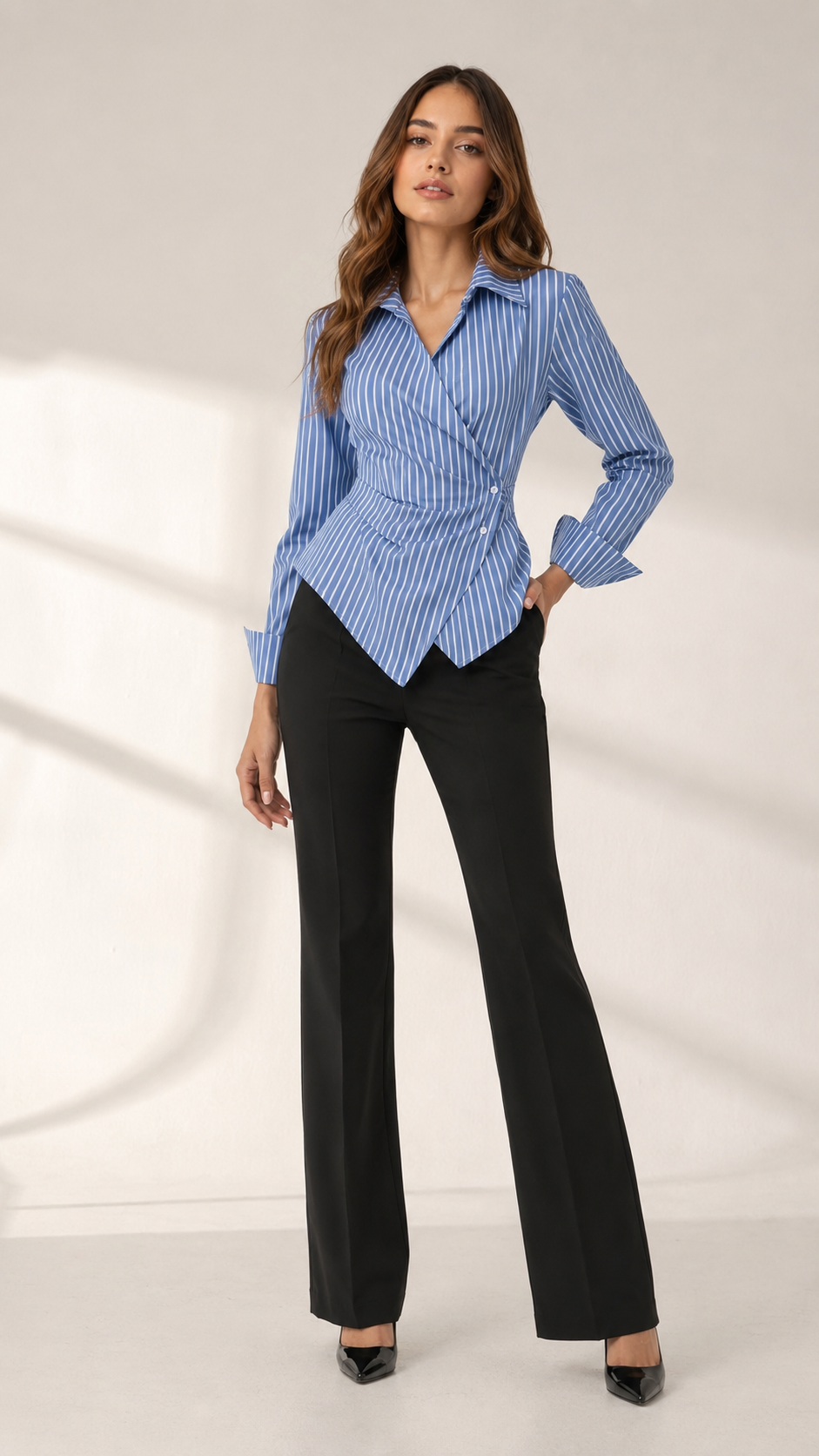 A woman modeling a blue and white vertically striped long-sleeve wrap blouse with black wide-leg trousers, standing against a light beige studio backdrop with soft shadow projections.