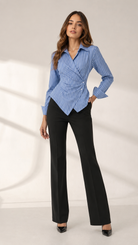 A woman modeling a blue and white vertically striped long-sleeve wrap blouse with black wide-leg trousers, standing against a light beige studio backdrop with soft shadow projections.