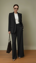 Woman in a black suit with a white top, holding a black handbag against a neutral background