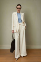 Woman in a cream suit with a blue top, holding a black handbag against a plain background